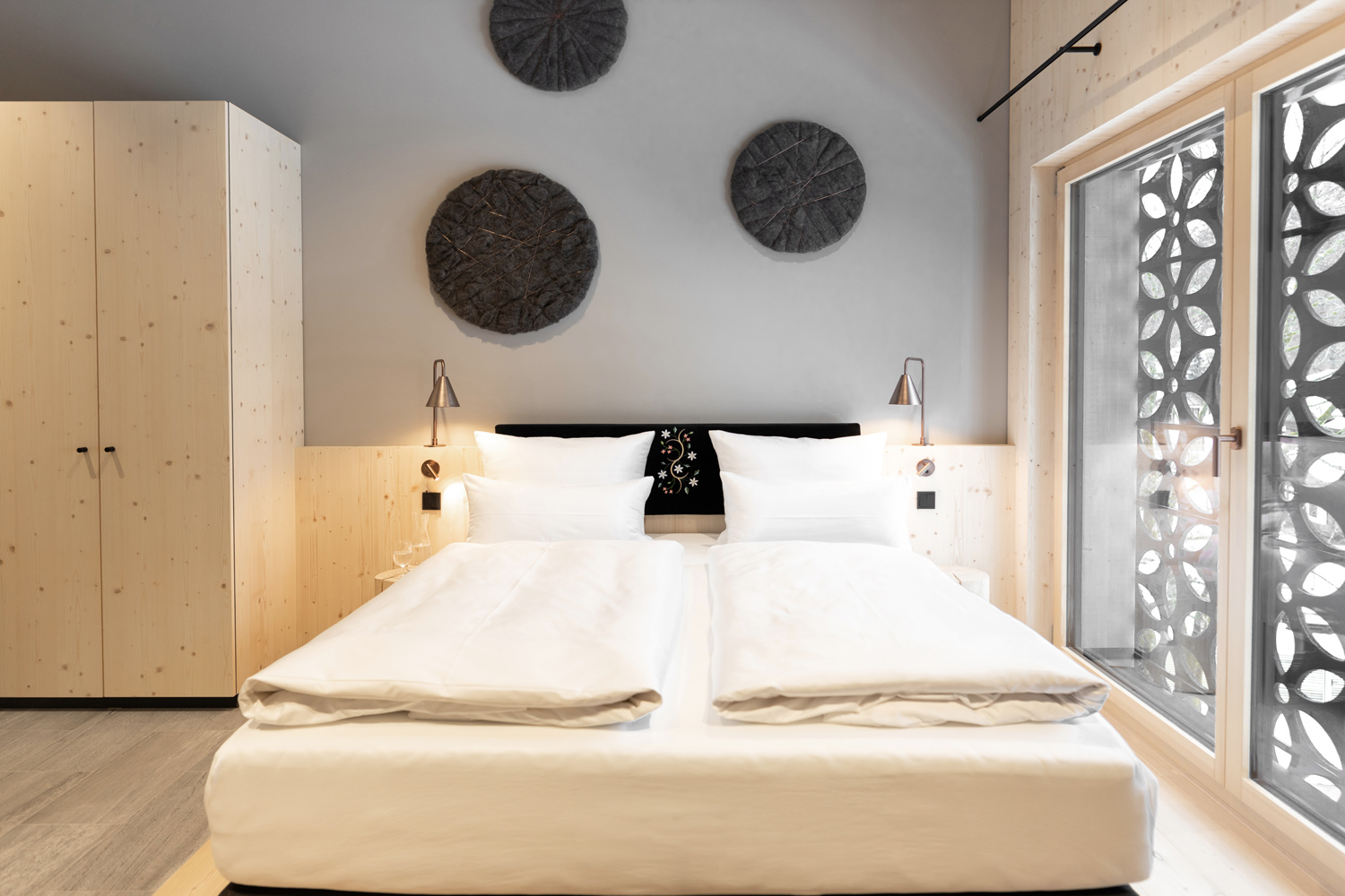 Gemütliches Bockspringbett für 2 Personen mit Nachtischlampen aus Kupfer und einem Schrank links vom Bett. Vor dem Fenster ist ein Rollo mit den Blumen Ornamenten